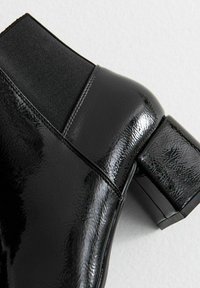 Botín negro de tobillo con un acabado brillante, paneles laterales elásticos y un tacón grueso en bloque. Textura suave, diseño moderno, costuras mínimas.