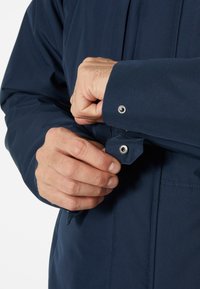 Chaqueta azul marino con una tela texturizada y detalles en los puños con botón a presión. Mano posicionada para abrochar el botón en la muñeca.