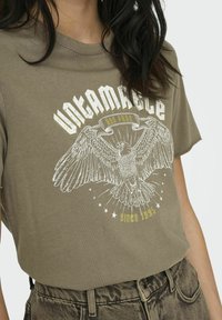 T-shirt en coton vert olive présentant un imprimé graphique d'un aigle avec des étoiles et le texte "Indomptable et Libre" en blanc et or. Manches courtes.