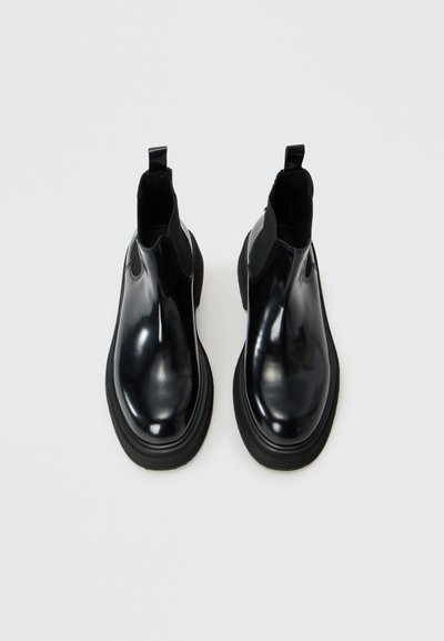 Bottines Chelsea noires brillantes avec un bout arrondi, des panneaux élastiques sur les côtés et des tirettes. Semelles en caoutchouc noir épaisses pour une meilleure adhérence.