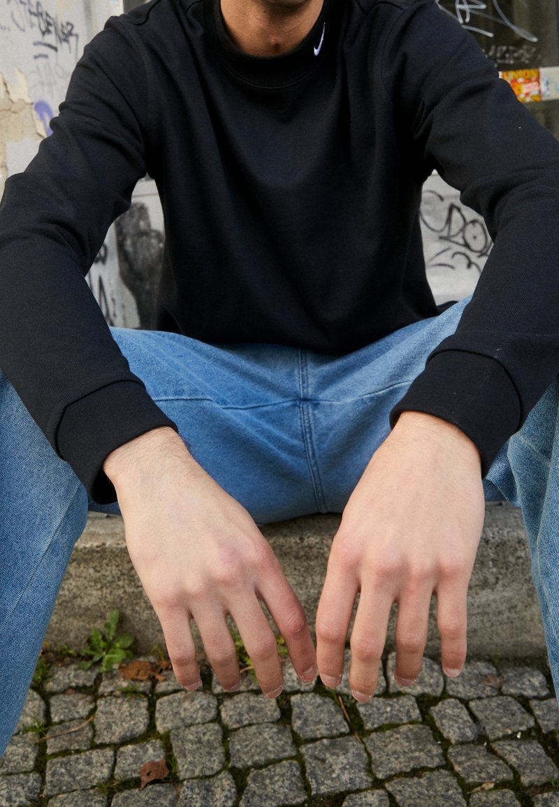 Personne portant une chemise noire à manches longues et un jean bleu, assise avec les mains reposant entre les genoux sur un pavé en pierre.