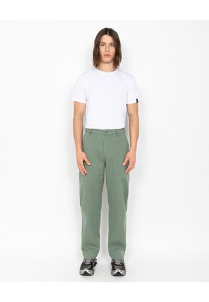 Jonge man die recht vooruit staat, gekleed in een eenvoudig wit T-shirt, groene rechte broeken en zwarte en grijze sneakers.