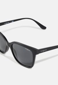 Ochelari de soare negri cu lentile închise, rama din plastic lucios și elemente de branding "vogue" argintii pe fiecare brată.