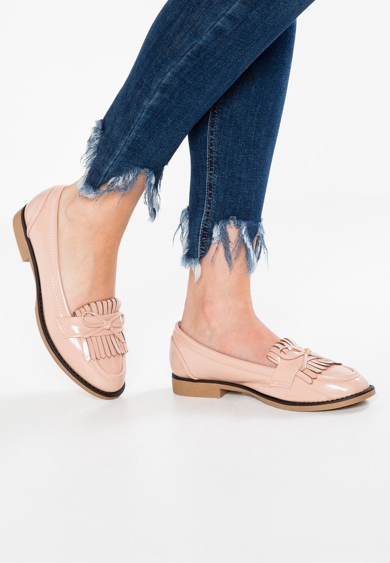 Mocassins en cuir verni rose clair avec un détail à franges et un nœud, dotés d'un talon bas et d'une texture lisse et brillante. Portés avec un jean bleu effiloché.