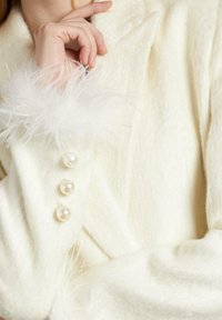 Chaqueta blanca de mezcla de lana que presenta botones de perla y un suave borde de plumas en los puños, mostrando una textura lisa y un diseño clásico.