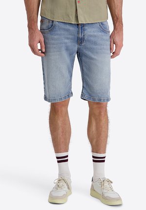 Mann trägt hellblaue Denim-Shorts, weiße Sneakers und weiße Socken mit zwei dunklen horizontalen Streifen und steht vor einem weißen Hintergrund.