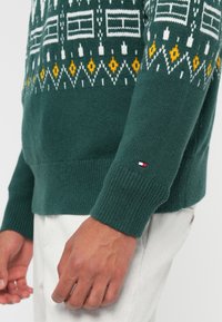 Green knit sweater features white and yellow geometric patterns, ribbed cuffs, and a small red, white, and blue logo on the sleeve.