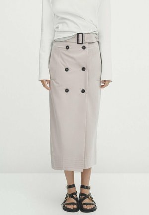 Femme portant une jupe midi beige boutonnée avec ceinture, un haut blanc à manches longues et des sandales plates noires à brides, debout devant un fond uni.