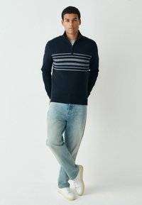 Marineblauer Strickpullover mit Reißverschlusskragen, versehen mit horizontalen hellblauen Streifen. Kombiniert mit hellblauen Jeans und weißen Sneakern.