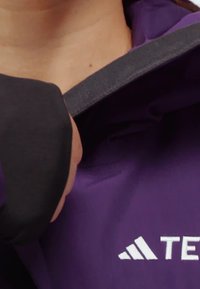 Hand, die einen grauen Riemen an einer lila Jacke mit weißem Adidas-Logo und teilweise sichtbarem "TEC"-Text auf dem Stoff anpasst.