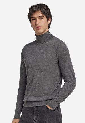 Junger Mann, der einen eng anliegenden grauen Rollkragenpullover und dunkle Jeans trägt, mit einer Hand in der Tasche vor einem schlichten weißen Hintergrund.