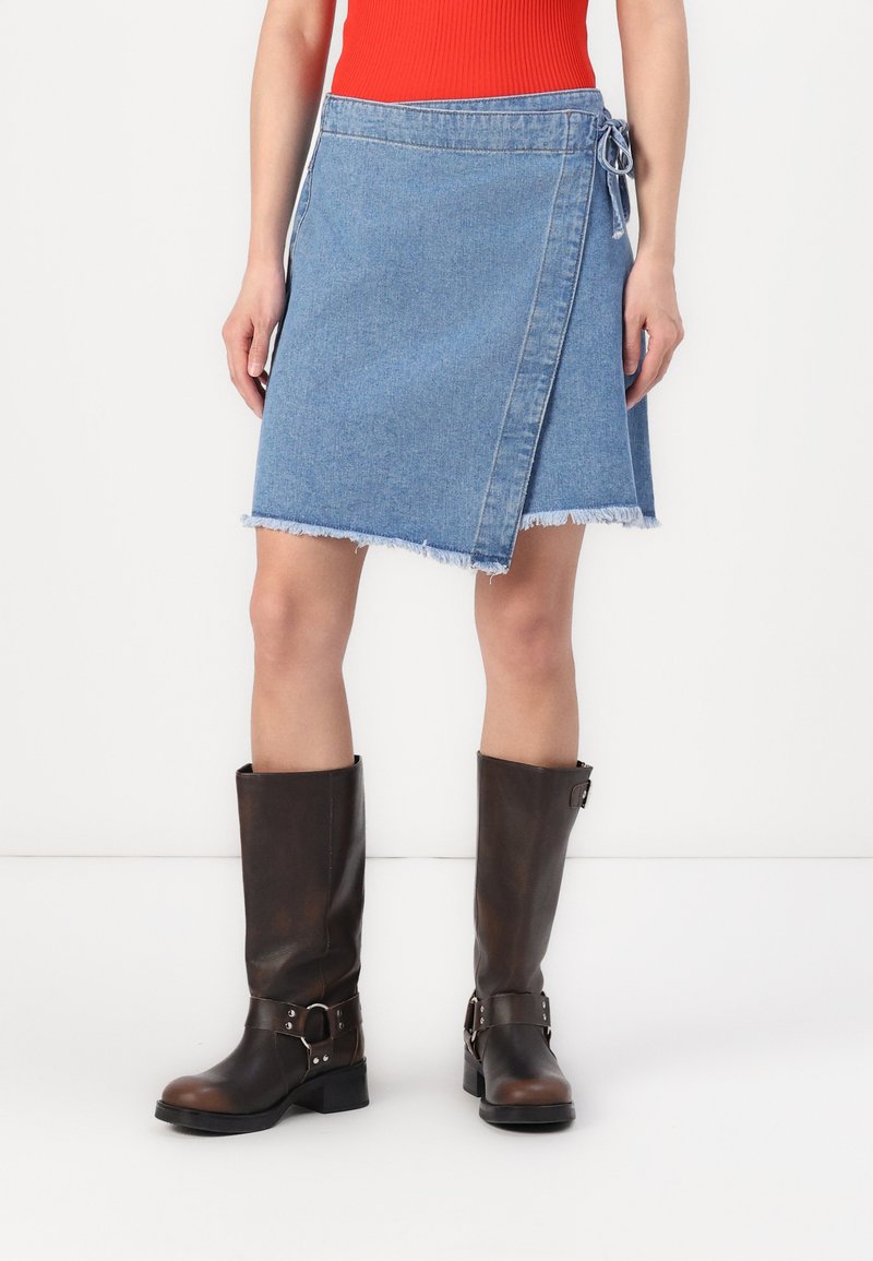 Denim omslagskjørt i lyseblått med frynset kant og sidesnor, kombinert med kne-høye brune lærstøvler med spenne-detaljer.