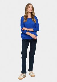 Blue knitted cardigan with round buttons, wide sleeves, and ribbed cuffs, paired with dark jeans and light sneakers with green accents.