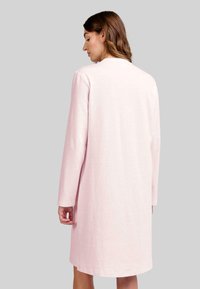 Mujer con cabello castaño claro vistiendo un vestido de manga larga, de color rosa claro, hasta la rodilla, de espaldas contra un fondo gris liso.