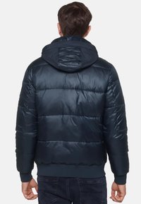 Mörkblå puffjacka med fast huva, quiltad design, ribbade ärmslut och nederkant. Mjuk, blanka yta; vadderad för isolering.