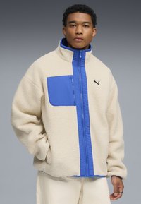 Fleece-takki vaalean kermanvärinen, sinisellä etupaneelilla ja kauluksella. Sisältää vetoketjullisen taskun rinnassa ja pienen mustan logon sivussa.
