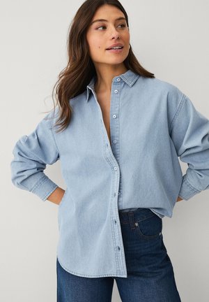 Kvinde med langt brunt hår iført en løs, lys denimskjorte, delvist knappet, kombineret med mørkeblå jeans, hænderne på hofterne, kigger mod højre.