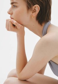 Pendiente de aro dorado en una persona con cabello corto. Piel lisa, camiseta gris y mano apoyada en la barbilla. Vista de perfil sobre un fondo liso.