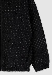 Giacca nera trapuntata con pois bianchi, dotata di zip frontale, polsini elasticizzati e un tessuto morbido e texturizzato.