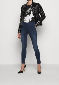 Chaqueta acolchada negra y brillante sobre una camiseta gráfica con un diseño blanco, combinada con jeans ajustados de color azul oscuro y tacones altos negros.