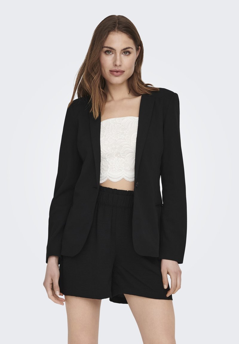 Blazer noir au design structuré, à manches longues, avec une fermeture à un bouton, superposé à un bandeau en dentelle blanche et à un short noir ample.