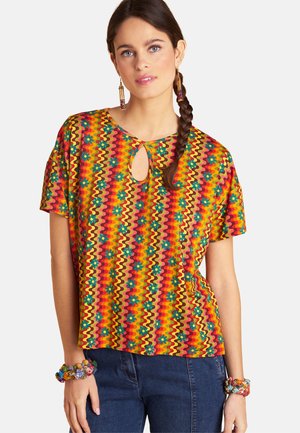 Frau trägt ein farbenfrohes Kurzarmoberteil mit wellenförmigen Streifen und Blumenmustern, kombiniert mit blauen Jeans und passenden Perlenarmbändern.