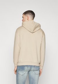 Beiger Hoodie mit großer Kapuze, lockerer Passform, gerippten Bündchen und Saum. Getragen mit hellblauen Jeans, die eine legere Rückansicht zeigen.