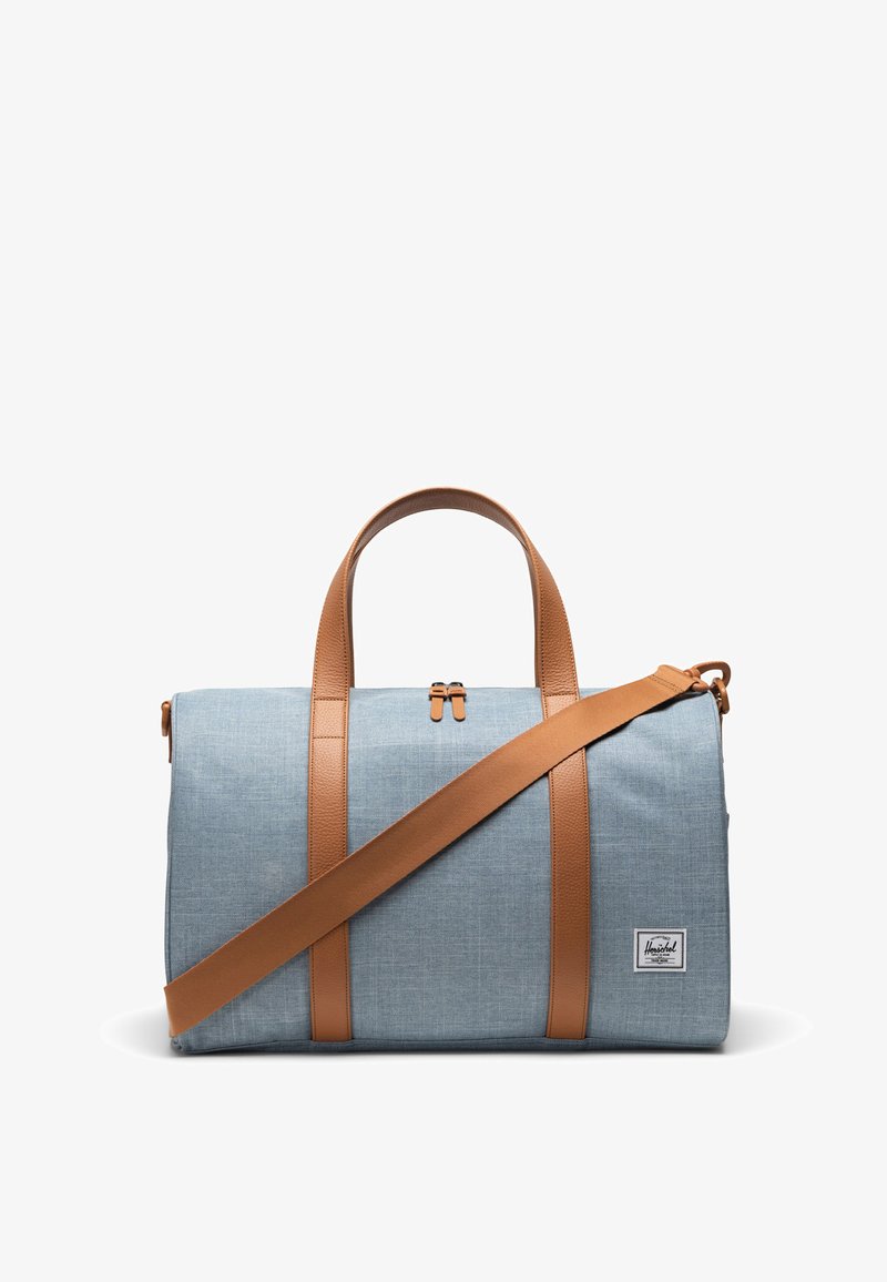 Cestovná taška svetlomodrej farby z látky s hnedými syntetickými koženými popruhmi, ktorá má horné zapínanie na zips a malú nášivku s logom značky.