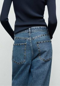 Personne portant un jean bleu délavé moyen avec des clous métalliques dessinant les poches et les coutures, associé à un haut noir ajusté à manches longues.