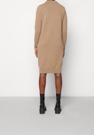 Woman standing with back to camera wearing a long-sleeve, knee-length beige sweater dress and black heeled ankle boots.