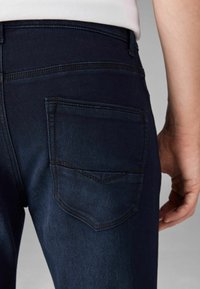 Jeans de mezclilla azul oscuro con un bolsillo trasero que presenta un sutil patrón de costura en zigzag y una textura suave, exhibidos en un ángulo.