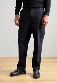 Pantalones cargo negros hechos de un tejido duradero, con bolsillos laterales, un corte relajado y combinados con zapatos negro de slip-on sobre un suelo de madera.