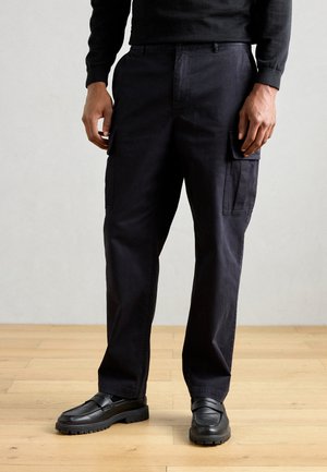 Black cargo pants made of durable fabric, featuring side pockets, a relaxed fit, and paired with black slip-on shoes on a wooden floor.
