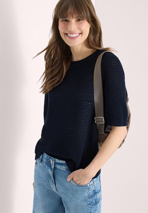 Femme souriante portant un haut noir en maille à manches courtes, un jean bleu clair et un sac à dos marron sur une épaule.