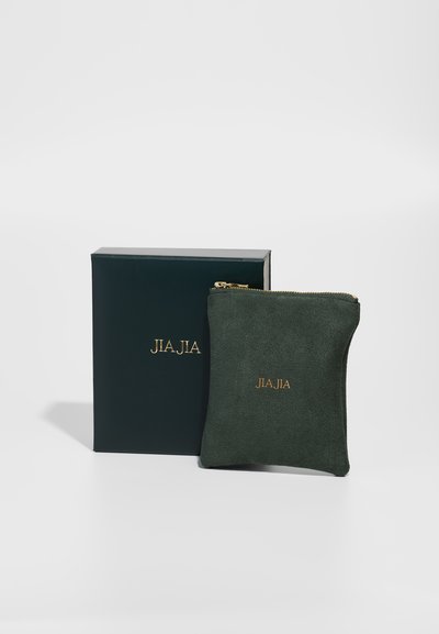Bolso de ante verde con el logo dorado "JIA JIA", acompañado de una caja a juego de color verde oscuro. El bolso cuenta con cierre de cremallera y una textura suave.