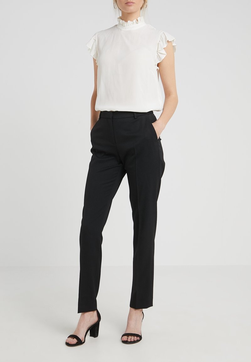 Femme portant un chemisier blanc sans manches avec des volants, un pantalon noir ajusté et des sandales noires à talon bloc ouvert, debout les mains dans les poches.