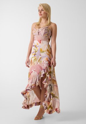 Femme debout portant une robe sans manches fleurie asymétrique avec des volants, tenant un sac à main beige et portant des sandales dorées à brides.