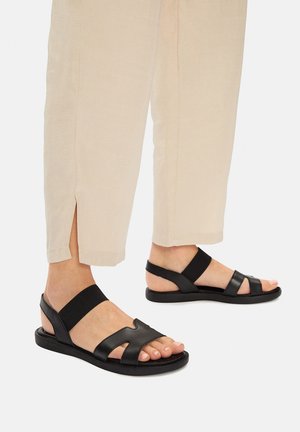 Sandalias negras de cuero con suela plana, con una correa elástica y un diseño recortado, usadas con pantalones cónicos beige claro.