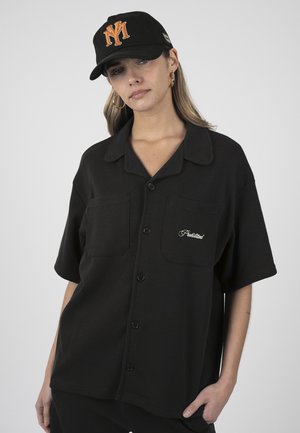 Giovane donna che indossa una camicia nera con bottoni con la scritta "Prohibited" e un berretto nero con il logo arancione "NY", in piedi su sfondo grigio.