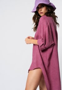 Vestito camicia in lino oversize di un intenso rosa profondo, con maniche lunghe e fessure laterali, abbinato a un cappello a secchiello tessuto di un leggero viola.