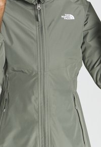 Wasserabweisende grüne Jacke mit Reißverschluss, elastischem Bund und Seitentaschen. Mit einem weißen Logo auf der linken Brust.