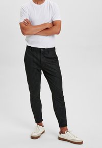 Slim-fit černé pruhované kalhoty s knoflíkovým zapínáním, kombinované s bílým tričkem s kulatým výstřihem a bílými teniskami s hnědými akcenty.