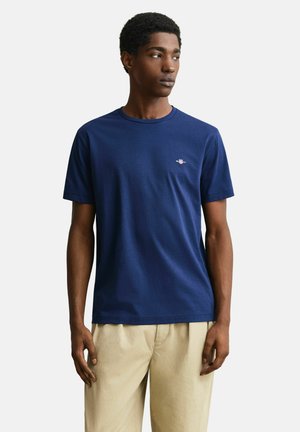 Jeune homme portant un T-shirt bleu marine à manches courtes et un pantalon beige, debout et regardant sur le côté sur un fond uni.