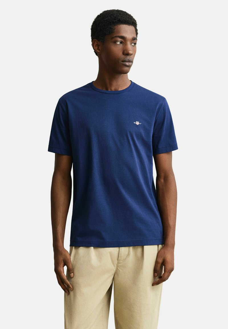 Jeune homme portant un T-shirt bleu marine à manches courtes et un pantalon beige, debout et regardant sur le côté sur un fond uni.