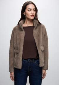 Faux fur jacket in light brown, zippered front, two patch pockets, and a soft texture. Worn over a brown top and dark blue jeans.