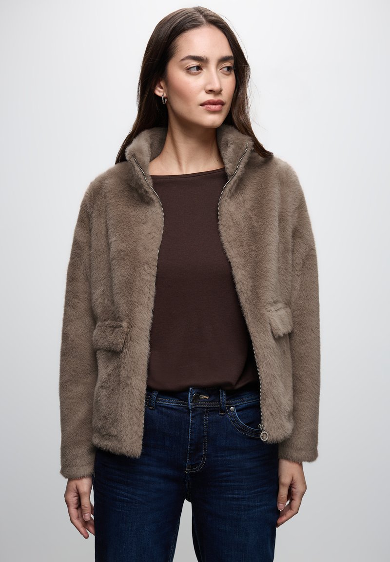 Faux fur jacket in light brown, zippered front, two patch pockets, and a soft texture. Worn over a brown top and dark blue jeans.
