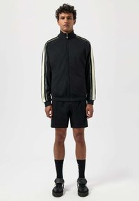Svart träningsjacka med hög krage och vita ränder på ärmarna, tillsammans med svarta shorts, svarta strumpor och svarta sneakers.