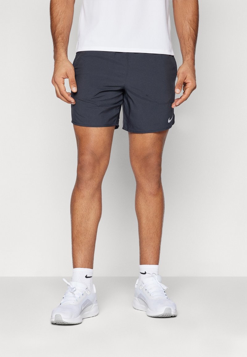 Homme portant un short de sport noir, des chaussettes blanches et des chaussures de course blanches, debout devant un fond uni.
