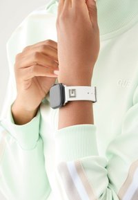 Reloj inteligente con una esfera cuadrada negra y una correa de silicona blanca que presenta una hebilla y un logotipo plateados. Lleva puesto en una muñeca, con una tela verde menta cercana.