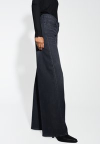 Jean en denim foncé à jambe large avec une taille haute, présentant une finition soignée et des détails de couture subtils. Associés à des bottes noires à bout pointu.